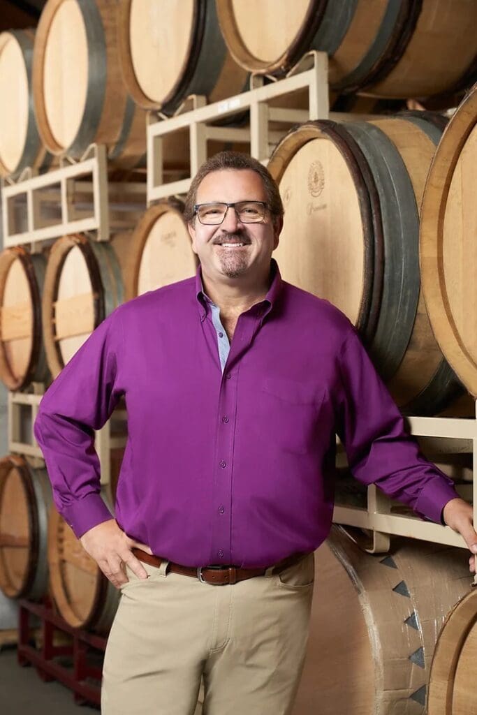 Man in a purple shirt smiling, standing in front of stacked wooden wine barrels in a winery.