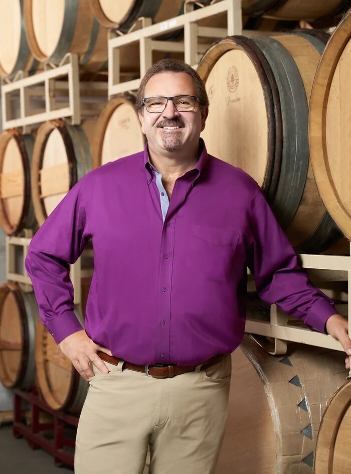 Man in a purple shirt smiling, standing in front of stacked wooden wine barrels in a winery.