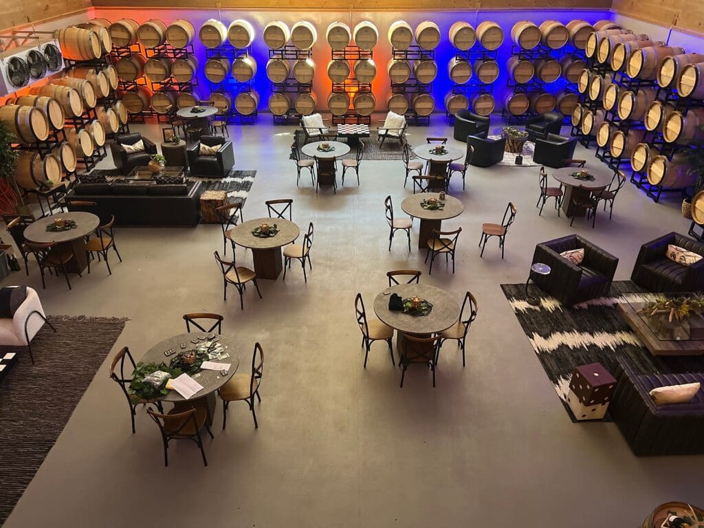 A spacious winery room with round tables, sofas, and wine barrels stacked along the walls, lit with colored lights.