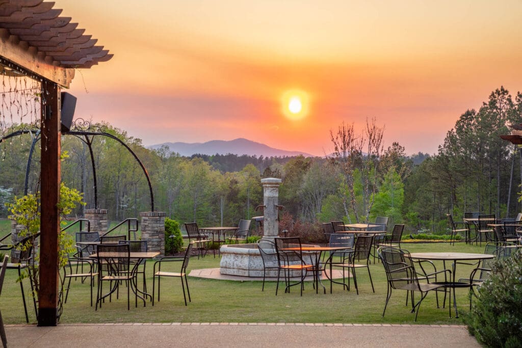 Outdoor patio with empty tables and chairs overlooking trees and mountains at sunset.