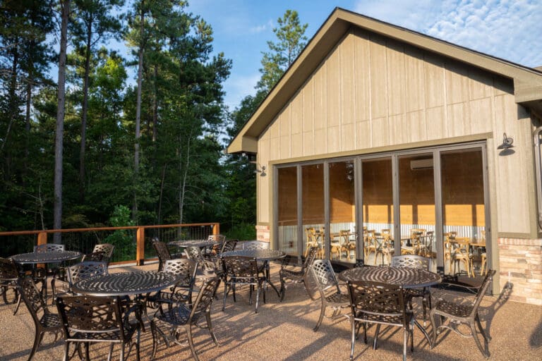 Outdoor patio with black metal tables and chairs beside a building, surrounded by trees under a blue sky.